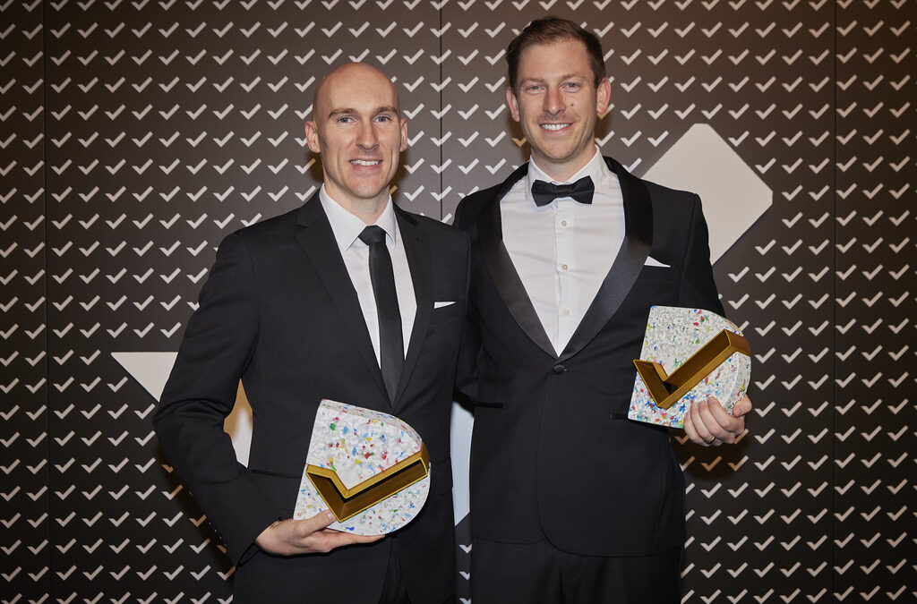 Two men in suits smile and hold two good design awards